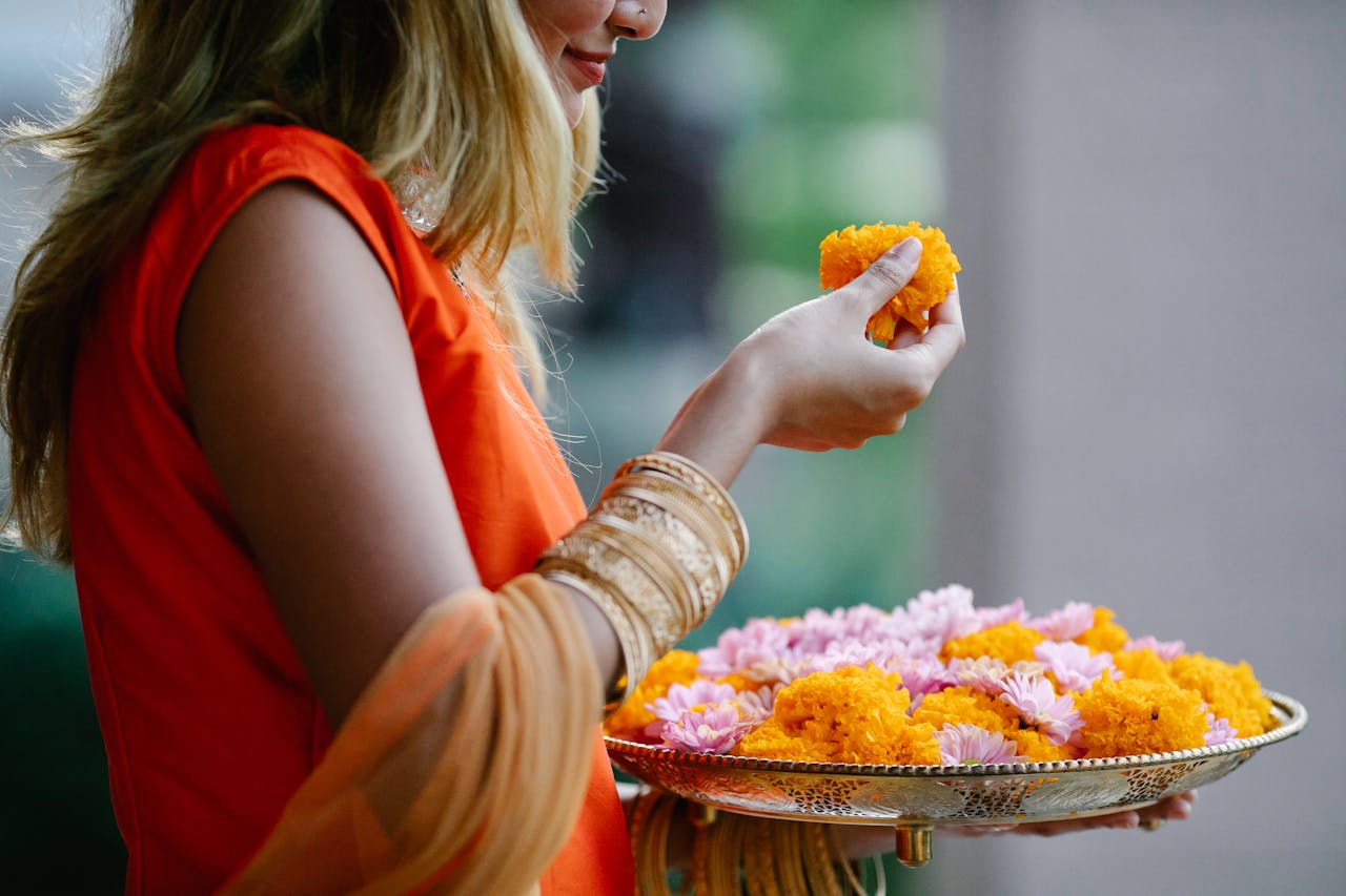 A woman in traditional attire holds a tray of marigold and chrysanthemum flowers.